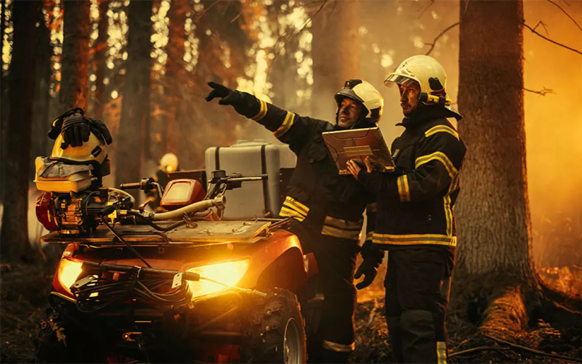 Two firefighters in a smoky forest look at a laptop and strategize next to an all-terrain vehicle laden with equipment.