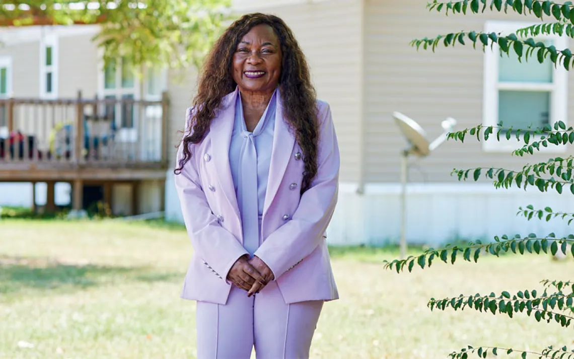 Catherine Coleman Flowers wears a lavender pantsuit and stands in front of a house, smiling at the camera.