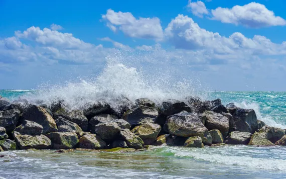 Waves crashing on a rock jetty in Dania Beach, Florida, with puffy clouds in the background