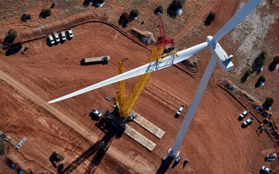 Aerial photo of a white wind turbine over a red dirt area, with a yellow crane attached and some cars scattered underneath