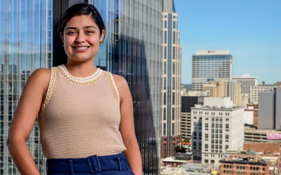 Laura Zapata stands against a glass wall inside a skyscraper with a city in the background