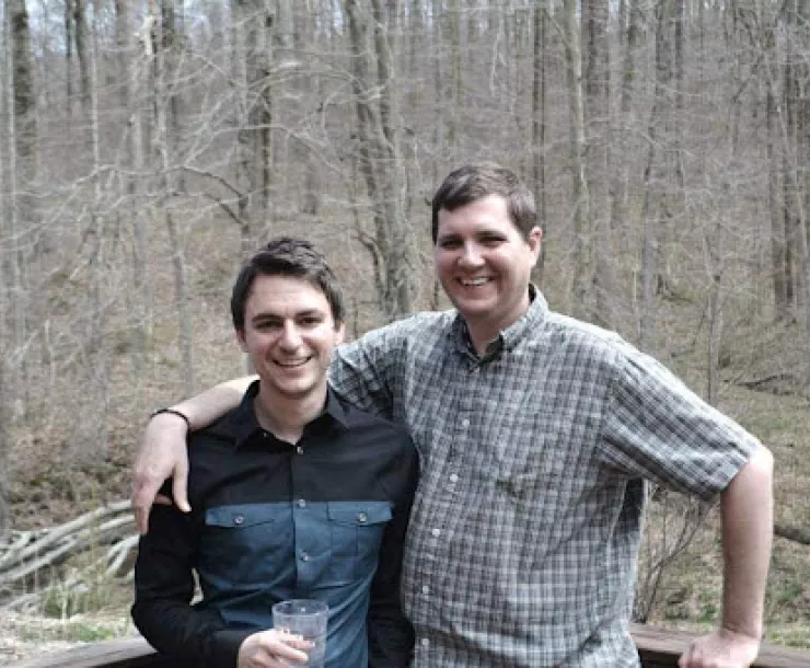 Two people stood with a forest backdrop. The taller person has their arm around the other. The trees are bare.