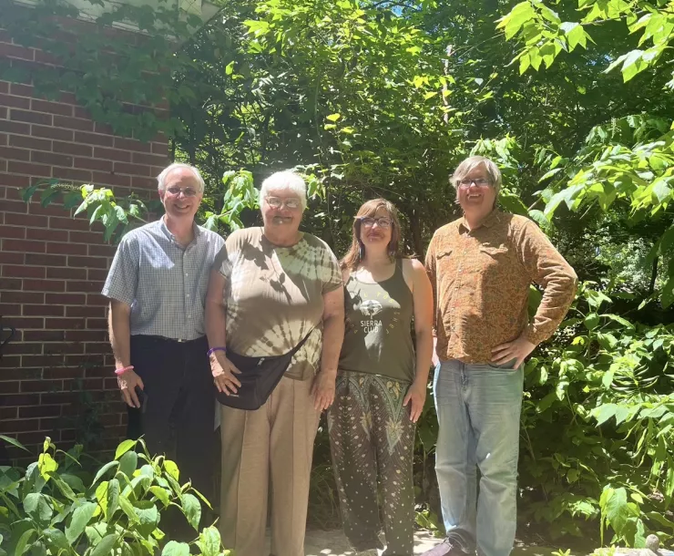Four people stand in a garden. The leaves on the trees are green and blue sky can be seen beyond the trees. The people are smiling and looking at the camera for the photo.