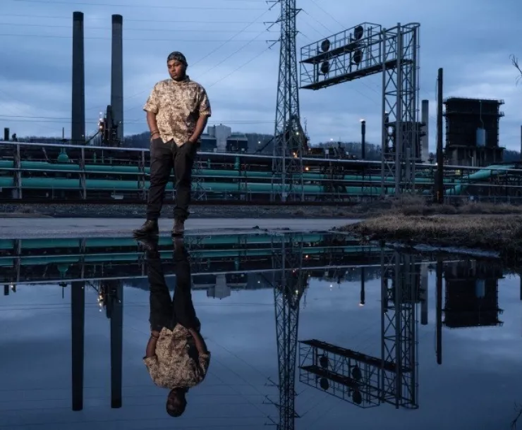 Qiyam Ansari stands with his hands in his pockets in front of an industrial complex and is reflected in a body of water.