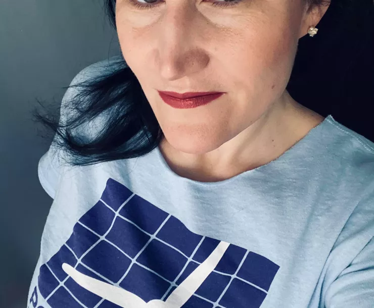 A close-up selfie of a woman with mid-length dark brown hair. She's wearing a blue t-shirt with part of a wind turbine showing