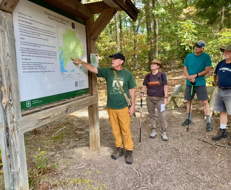 Four people at a trailhead looking at a map of the park. A couple of the folks have walking poles and they're all wearing hats to protect from the sun and bugs. The ground has some fallen brown leaves but the leaves on the trees behind them are green.