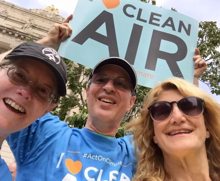 alt="Three people at a rally. The person in the middle is holding up a protest sign saying Clean Air in black letters on a turquoise background. He is also wearing a blue t-shirt that says #actonclimate. He and the two people either side of him are smiling for the photo."