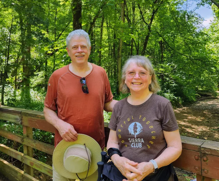 Two people stood next to each other on a trail. It's a sunny day and the trees have green leaves. They are smiling. One is wearing a Sierra Club t-shirt that says Powerful Together.