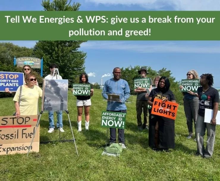 Image of a group of people of many ages and races standing in a park holding protest signs about affordable energy and wanting clean energy. Two large smoke stacks are in the background.