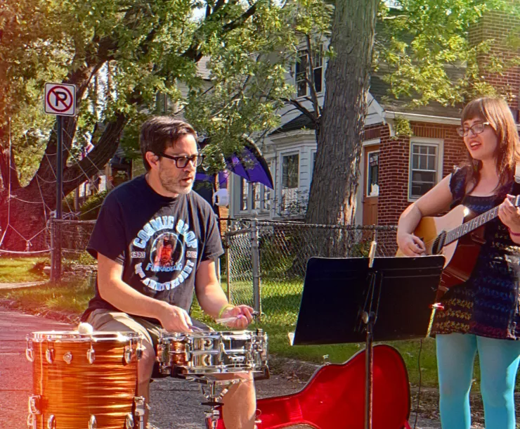 Two people playing music in the street. One is sitting down playing drums, the other is standing playing acoustic guitar. They are both singing. It's a sunny day and some trees are visible in the yards in front of the houses behind them.