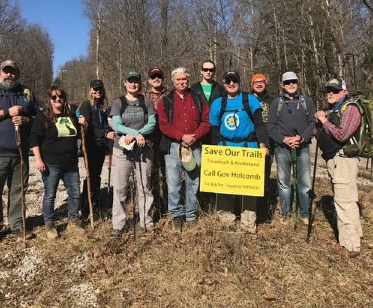 A group of eleven people at a trailhead. Some of them having hiking poles. The sky is blue and the trees are bare. Some of the people are wearing caps and hiking backpacks.