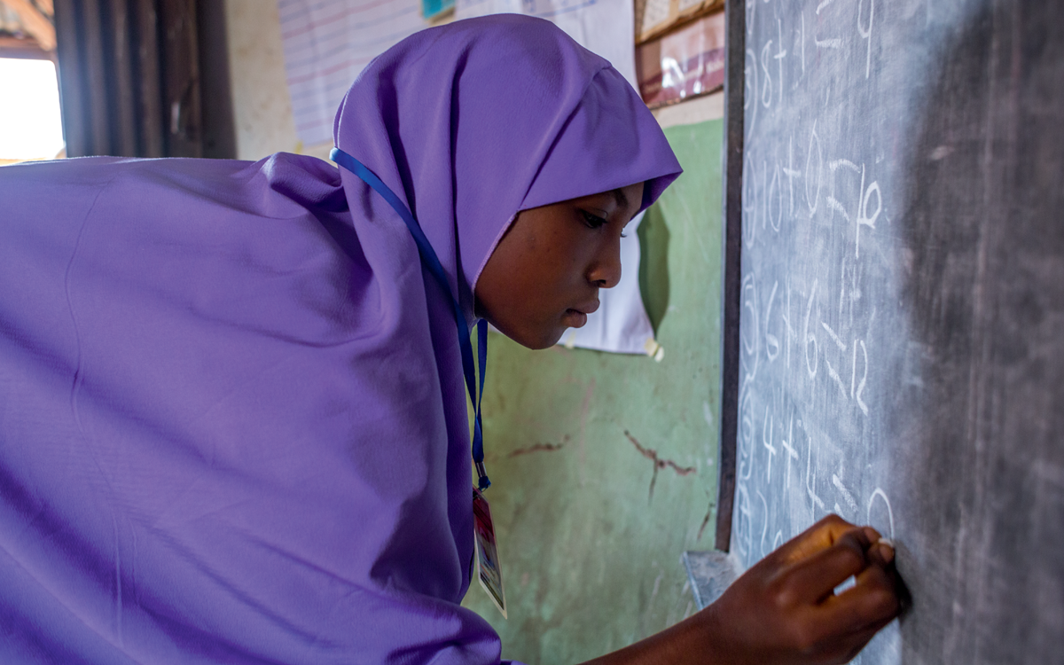 A teen girl in a purple hijab is bending over and writing on a chalkboard during a numeracy class in an out of school safe space.