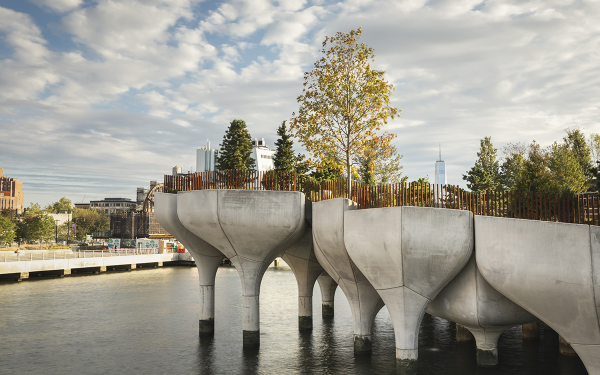 A tree-filled park balanced over the water on what look like giant concrete golf tees, with skyscrapers and water in background. 