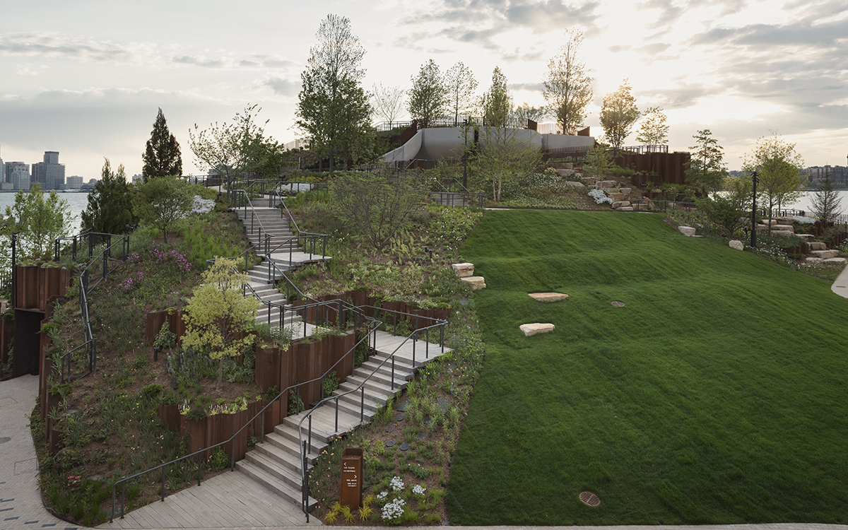 Hill of park greenery with switchback of staircases and a view in the distance of skyscrapers behind the water. 